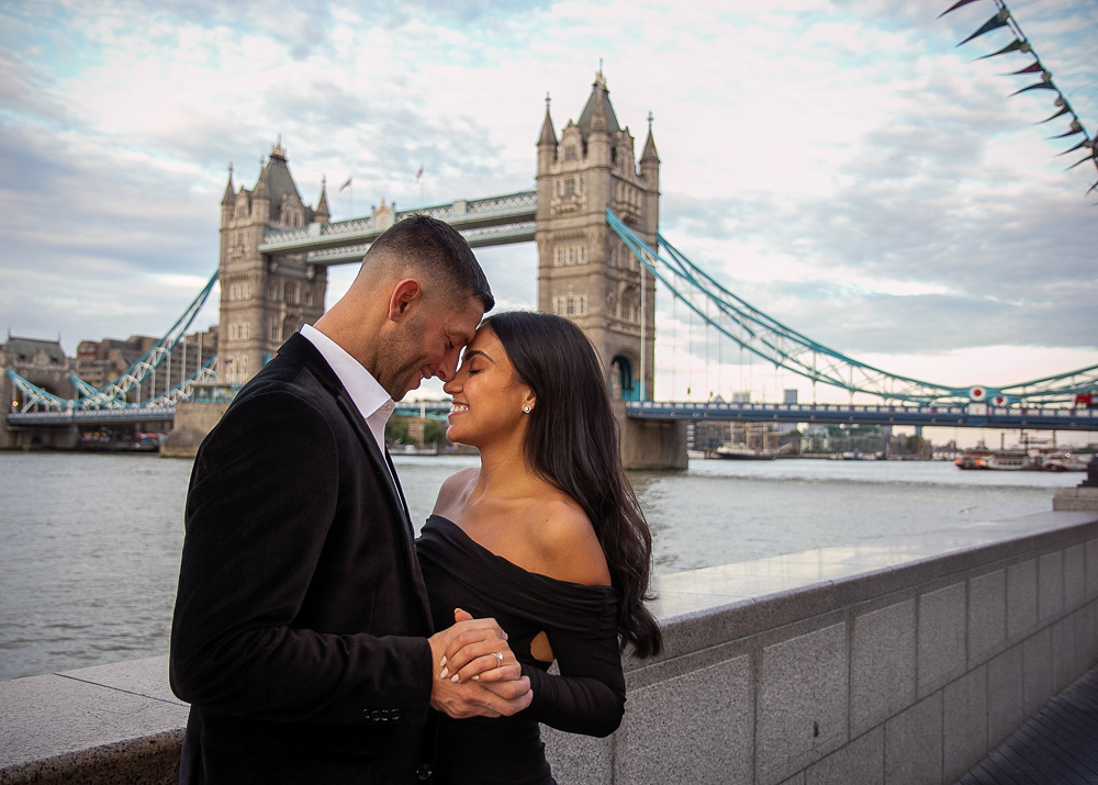 Newly engaged couple dressed in black posing for engagement photos in front of the Tower Bridge in London
