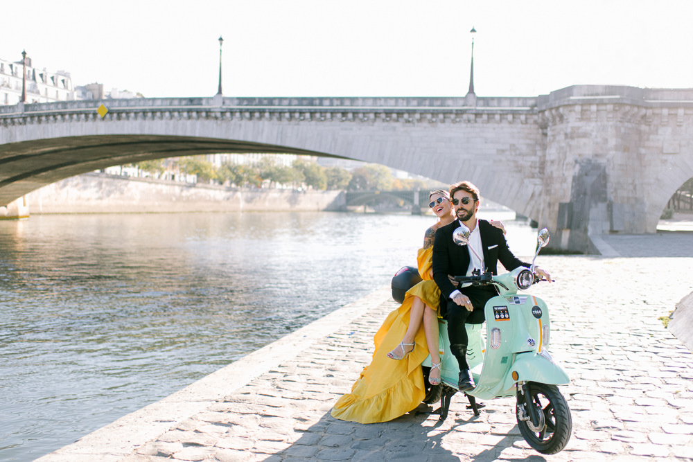 Couple wearing yellow dress and dark suit posing for couples photos on a vespa in Paris, France