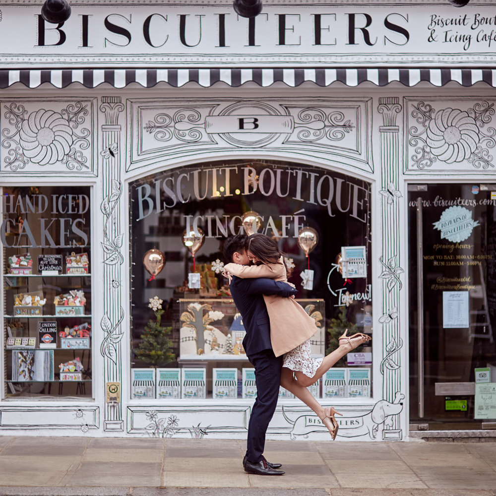 Guy picking his fiancée up while posing for engagement photos in front of a biscuits shop in Nothing Hill