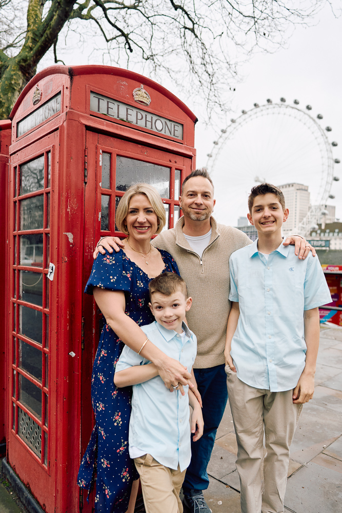 Happy family posing for pictures in front of London Eye, next to a red telephone boot