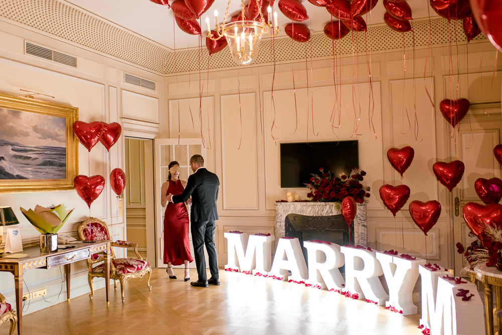 Hotel room proposal - intimate setting with red heart balloons and marry me letters