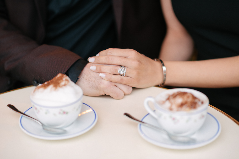 Newly engaged couple holding hands next to cappuccino cups during professional engagement photoshoot - The Now Time