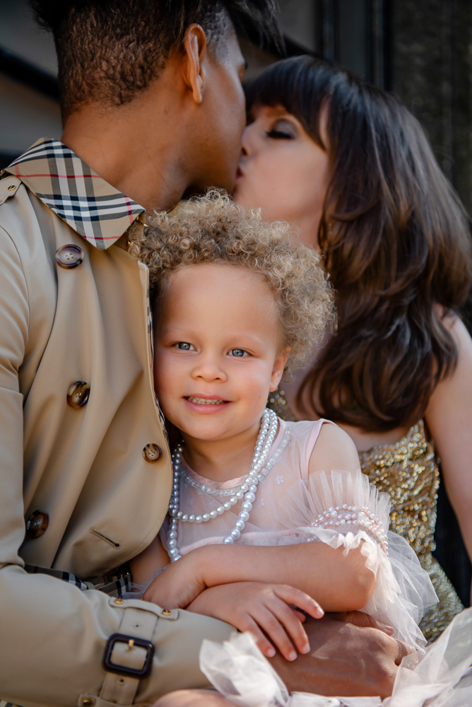 Beautigul girl with blue eyes smiling while parents kissing on photo shoot by London family photographer the now time