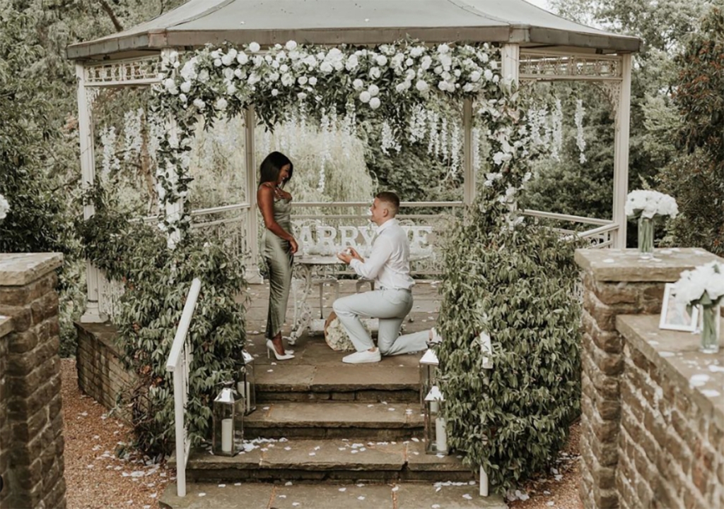 Man dressed in white, asking his girlfriend to marry him under a pergola filled with white flowers